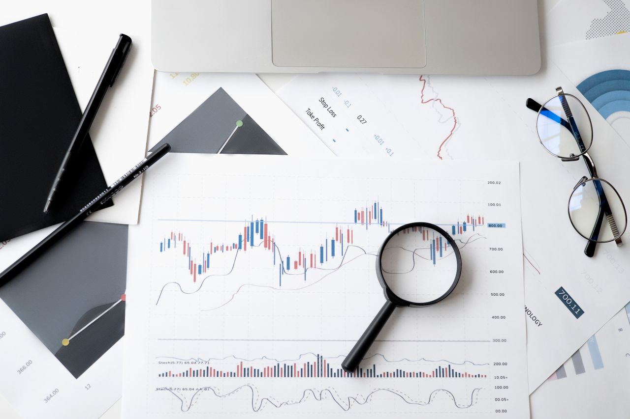 contact-img Flat lay of stock market analysis documents with magnifying glass, pens, and glasses.